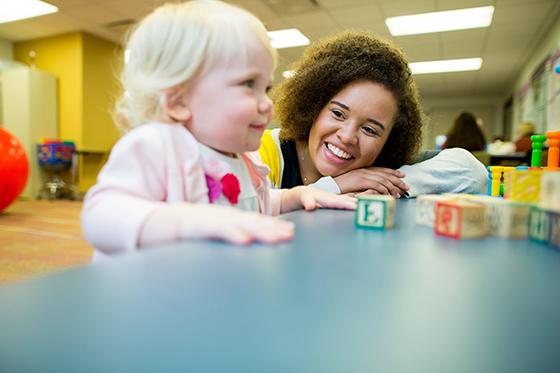 图为一名英国波胆网站的学生趴在桌子上，对着一个正在玩字母积木的小孩微笑