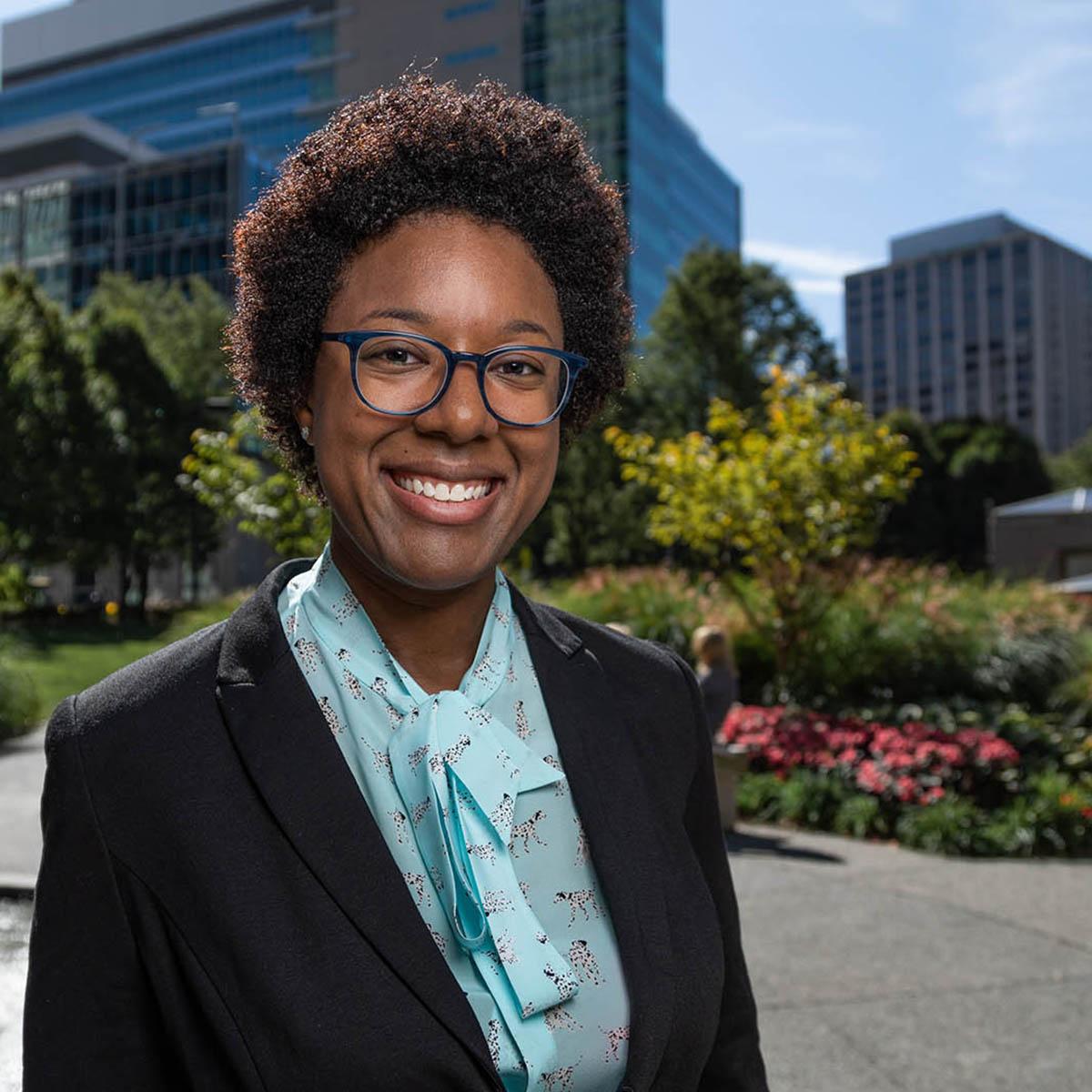 Photo of a smiling Black woman in professional clothes, posing in front of buildings and greenery in downtown Pittsburgh