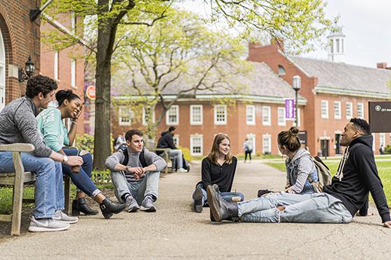 Photo of a group of six Chatham University students sitting outside a red brick academic building on the ground and benches talking